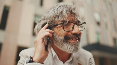 Gri ceketli, gözlüklü, sakallı, olgun iş adamı cep telefonu kullanıyor. Orta yaşlı yönetici ofisin dışında otururken akıllı telefondan konuşuyor.