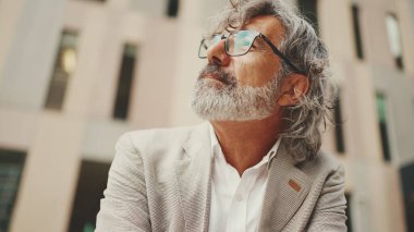 Gözlüklü, gri ceketli, sakallı, olgun bir işadamı cep telefonunu elinde tutuyor ve etrafa bakıyor.