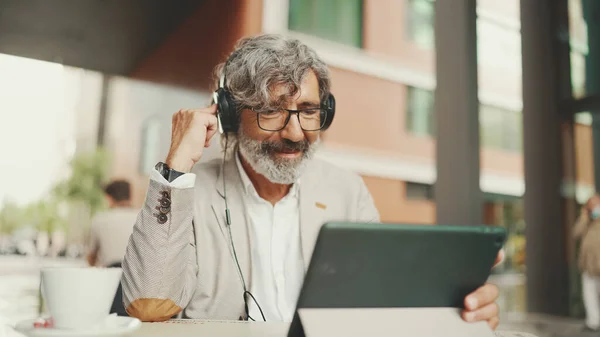 Mature businessman with beard in eyeglasses and headphones, sits in an outdoor cafe and works using tablet