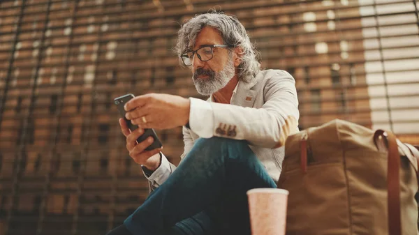 Mature businessman with beard in eyeglasses wearing gray jacket is using cell phone. Middle aged manager scrolling information on his smartphone while sitting outside the office
