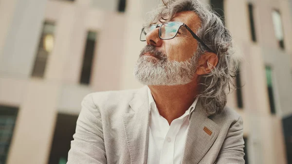 Mature businessman with beard in eyeglasses wearing gray jacket holds cellphone in his hand and looks around pondering