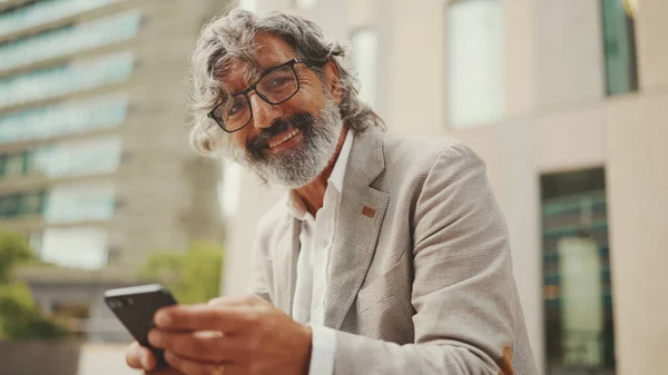 Mature businessman with beard in eyeglasses wearing gray jacket holds cellphone in his hand and looks around pondering