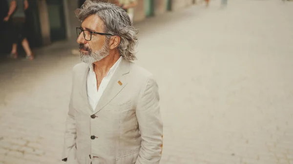 Mature businessman with beard in eyeglasses wearing gray jacket walks down the street past modern buildings