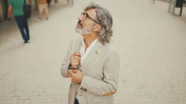 Mature businessman with beard in eyeglasses wearing gray jacket walks down the street past modern buildings