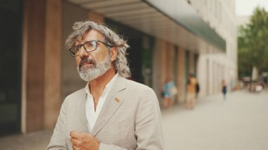 Gözlüklü, gri ceketli, olgun bir iş adamı modern binaların önünden geçiyor.