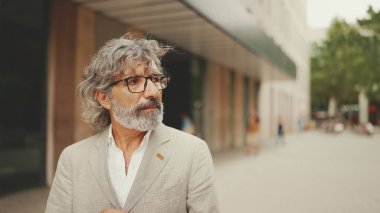 Gözlüklü, gri ceketli, olgun bir iş adamı modern binaların önünden geçiyor.