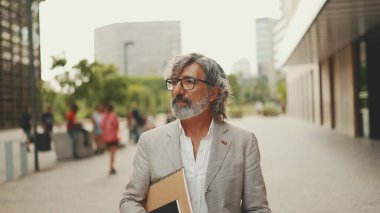 Gözlüklü, sakallı, gri ceketli, gülümseyen olgun bir işadamı etrafına bakıyor ve modern binaların önünden geçiyor.