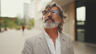 Gözlüklü, gri ceketli, sakallı, olgun bir işadamı gibi modern binaların önünden geçeceğiz.
