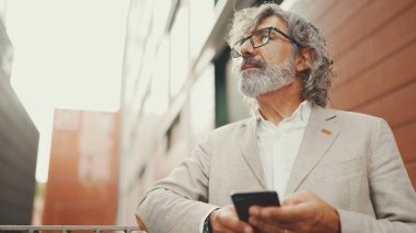 Gözlüklü, sakallı, olgun bir iş adamı iş merkezinin arka planında caddede duruyor. Orta yaşlı yönetici akıllı telefona mesaj iletisi kaydırıyor.