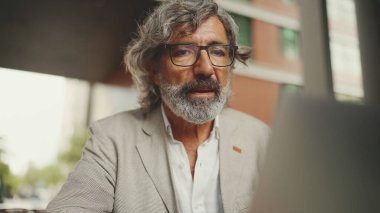 Gri ceketli, gözlüklü, sakallı, olgun iş adamı kafede oturuyor. Bilgisayarda orta yaşlı yönetici başarılı adam kullanıcısı açık kafede oturuyor
