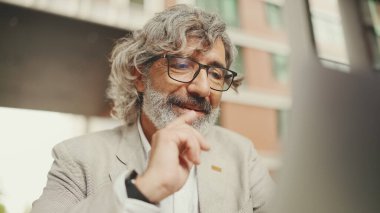 Gözlüklü, gri ceketli, sakallı, olgun bir işadamı kafede oturuyor. Orta yaşlı yönetici video konferansı yapıyor, bilgisayarda video görüşmesi yapıyor açık kafede oturuyor.
