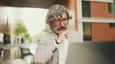 Gri ceketli, gözlüklü, sakallı, olgun iş adamı kafede oturuyor. Bilgisayarda orta yaşlı yönetici başarılı adam kullanıcısı açık kafede oturuyor