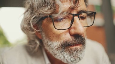 Gözlüklü, sakallı, gri ceketli, laptoplu, bilgisayarlı açık kafede oturan yetişkin iş adamı.