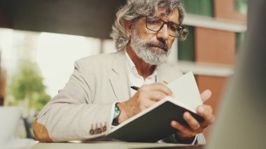 Gözlüklü, sakallı, dizüstü bilgisayarlı ve notlar alan, açık kafede oturan olgun bir iş adamı. Başarılı bir adam doğadaki kafede oturuyor. Mobil ofis çalışanı.