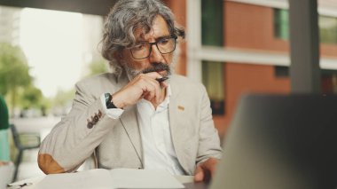 Gözlüklü, sakallı, dizüstü bilgisayarlı ve notlar alan, açık kafede oturan olgun bir iş adamı. Başarılı bir adam doğadaki kafede oturuyor. Mobil ofis çalışanı.
