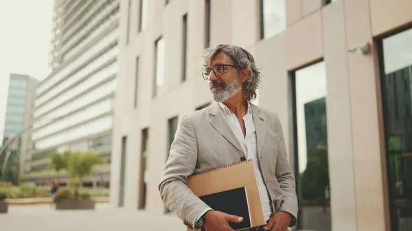 Smiling mature businessman with beard in eyeglasses wearing gray jacket looks around and walks down the street past modern buildings