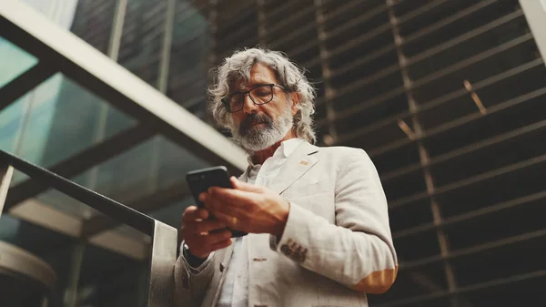 Mature businessman with beard in glasses wearing gray jacket is standing on the street at the business center background. Middle aged manager texting message scrolling tapping to smartphone