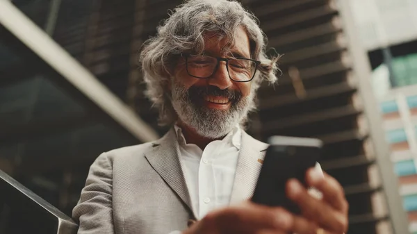 Mature businessman with beard in glasses stands on the street at the business center background. Middle aged manager texting message scrolling tapping to smartphone. Joy from good deal