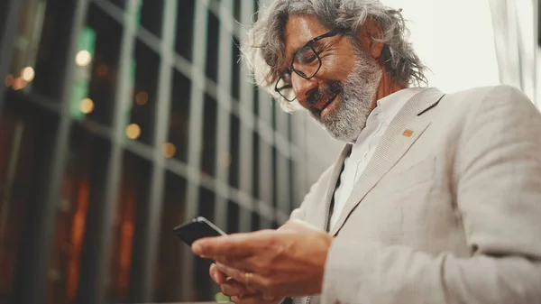 Mature businessman with beard in glasses stands on the street at the business center background. Middle aged manager texting message scrolling tapping to smartphone