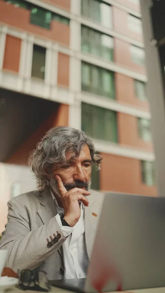 Clouse up, mature businessman with beard in eyeglasses wearing gray jacket sits on cafe. Middle aged manager successful man user on laptop pc computer sit at cafe outdoors