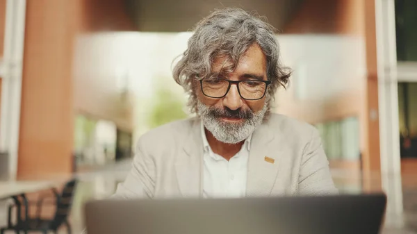 Mature businessman with beard in eyeglasses wearing gray jacket sits in cafe. Middle aged manager having video conference, video call on laptop pc computer sit at cafe outdoor