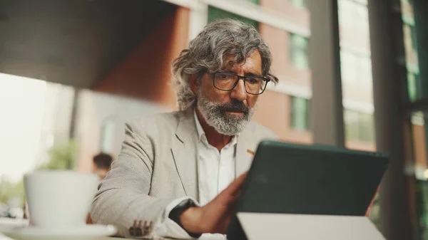 Mature businessman with beard in eyeglasses wearing gray jacket, working on tablet, sitting in an outdoor cafe. Successful man sitting at table in cafe in nature Mobile office freelancer