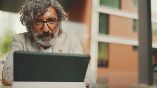 Mature businessman with beard in eyeglasses wearing gray jacket, working on tablet, sitting in an outdoor cafe. Successful man sitting at table in cafe in nature Mobile office freelancer