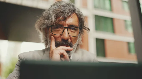 Mature businessman with beard in eyeglasses wearing gray jacket, working on tablet, sitting in an outdoor cafe. Successful man sitting at table in cafe in nature Mobile office freelancer