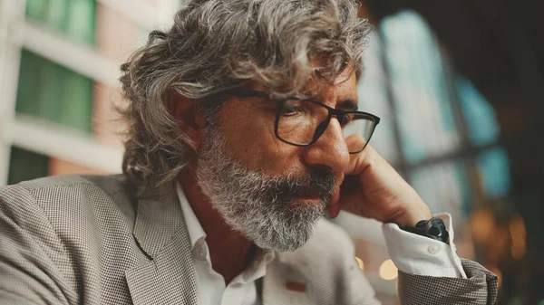 Mature businessman with beard in eyeglasses wearing gray jacket, working on tablet, sitting in an outdoor cafe. Successful man sitting at table in cafe in nature Mobile office freelancer
