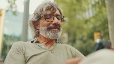 Gri saçlı, sakallı, sıradan kıyafetler giyen orta yaşlı bir adam bankta oturuyor ve cep telefonu kullanıyor. Gözlüklü olgun bir beyefendi akıllı telefondan mesaj yazar.