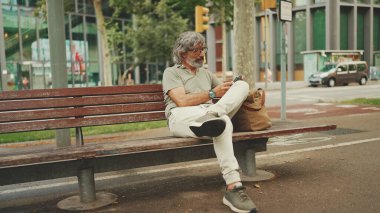 Gri saçlı, sakallı, sıradan kıyafetler giyen orta yaşlı bir adam bankta oturuyor ve cep telefonu kullanıyor. Gözlüklü olgun bir beyefendi akıllı telefondan mesaj yazar.