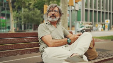 Gri saçlı, sakallı, sıradan kıyafetler giyen orta yaşlı bir adam bankta oturuyor ve cep telefonu kullanıyor. Gözlüklü olgun bir beyefendi akıllı telefondan mesaj yazar.