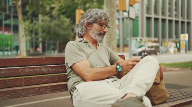 Gri saçlı, sakallı, sıradan kıyafetler giyen orta yaşlı bir adam bankta oturuyor ve cep telefonu kullanıyor. Gözlüklü olgun bir beyefendi akıllı telefondan mesaj yazar.