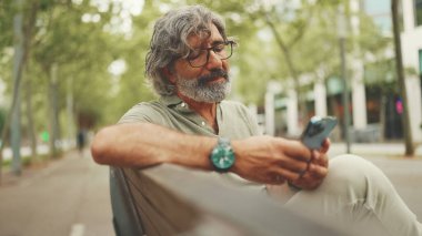 Gri saçlı, sakallı, sıradan kıyafetler giyen orta yaşlı bir adam bankta oturuyor ve cep telefonu kullanıyor. Gözlüklü olgun bir beyefendi akıllı telefondan mesaj yazar.