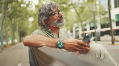 Gri saçlı, sakallı, sıradan kıyafetler giyen orta yaşlı bir adam bankta oturuyor ve cep telefonu kullanıyor. Gözlüklü olgun bir beyefendi akıllı telefondan mesaj yazar.