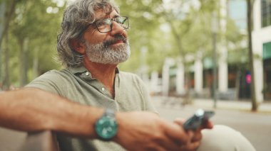 Gri saçlı, sakallı, sıradan kıyafetler giyen orta yaşlı bir adam bankta oturuyor ve cep telefonu kullanıyor. Gözlüklü olgun bir beyefendi akıllı telefondan mesaj yazar.
