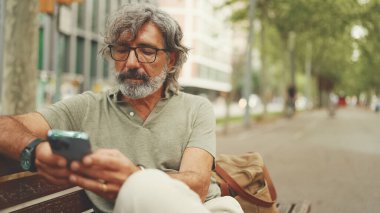 Gri saçlı, sakallı, sıradan kıyafetler giyen orta yaşlı bir adam bankta oturuyor ve cep telefonu kullanıyor. Gözlüklü olgun bir beyefendi akıllı telefondan mesaj yazar.