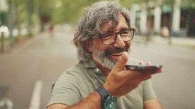 Gri saçlı, sakallı, sırıtan orta yaşlı adam bankta oturuyor ve cep telefonu kullanıyor. Gözlüklü olgun bir beyefendi hoparlörden ses tanıma mesajı kaydediyor.