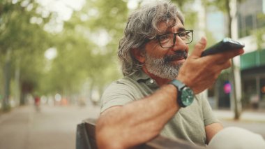 Gri saçlı, sakallı, sırıtan orta yaşlı adam bankta oturuyor ve cep telefonu kullanıyor. Gözlüklü olgun bir beyefendi hoparlörden ses tanıma mesajı kaydediyor.