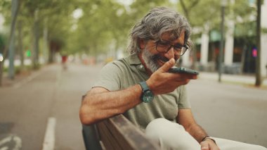Gri saçlı, sakallı, sırıtan orta yaşlı adam bankta oturuyor ve cep telefonu kullanıyor. Gözlüklü olgun bir beyefendi hoparlörden ses tanıma mesajı kaydediyor.