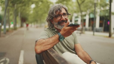 Gri saçlı, sakallı, sırıtan orta yaşlı adam bankta oturuyor ve cep telefonu kullanıyor. Gözlüklü olgun bir beyefendi hoparlörden ses tanıma mesajı kaydediyor.