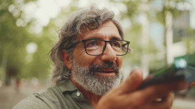 Gri saçlı, sakallı, sırıtan orta yaşlı adam bankta oturuyor ve cep telefonu kullanıyor. Gözlüklü olgun bir beyefendi hoparlörden ses tanıma mesajı kaydediyor.