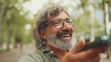 Gri saçlı, sakallı, sırıtan orta yaşlı adam bankta oturuyor ve cep telefonu kullanıyor. Gözlüklü olgun bir beyefendi hoparlörden ses tanıma mesajı kaydediyor.