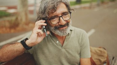 Gri saçlı ve sakallı, sıradan kıyafetler giyen orta yaşlı bir adam bankta oturuyor ve cep telefonu kullanıyor. Gözlüklü, akıllı telefondan konuşan olgun bir beyefendi.