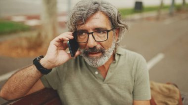 Gri saçlı ve sakallı, sıradan kıyafetler giyen orta yaşlı bir adam bankta oturuyor ve cep telefonu kullanıyor. Gözlüklü, akıllı telefondan konuşan olgun bir beyefendi.