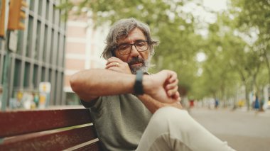 Gri saçlı ve sakallı, sıradan kıyafetler giyen orta yaşlı bir adam bankta oturuyor ve cep telefonu kullanıyor. Gözlüklü, akıllı telefondan konuşan olgun bir beyefendi.