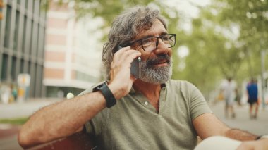 Gri saçlı ve sakallı, sıradan kıyafetler giyen orta yaşlı bir adam bankta oturuyor ve cep telefonu kullanıyor. Gözlüklü, akıllı telefondan konuşan olgun bir beyefendi.