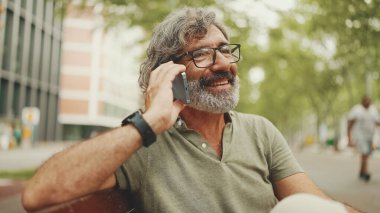 Gri saçlı ve sakallı, sıradan kıyafetler giyen orta yaşlı bir adam bankta oturuyor ve cep telefonu kullanıyor. Gözlüklü, akıllı telefondan konuşan olgun bir beyefendi.