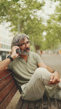 Gri saçlı ve sakallı, sıradan kıyafetler giyen orta yaşlı bir adam bankta oturuyor ve cep telefonu kullanıyor. Gözlüklü, akıllı telefondan konuşan olgun bir beyefendi.
