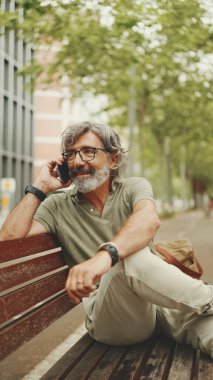 Gri saçlı ve sakallı, sıradan kıyafetler giyen orta yaşlı bir adam bankta oturuyor ve cep telefonu kullanıyor. Gözlüklü, akıllı telefondan konuşan olgun bir beyefendi.
